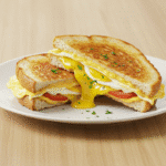 Grillowany tost z jajkiem i serem, pokrojony na pół, z roztopionym serem i świeżymi dodatkami.