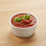 Sos do pizzy w małej miseczce, składający się z pomidorów, czosnku, bazylii i oliwy z oliwek, z przyprawami rozłożonymi obok.